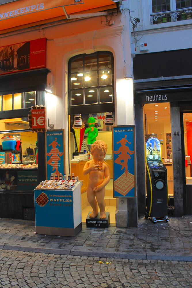 !Postać o kształcie słynnej figurki Manneken pis witająca turystów przed budką z goframi w Brukseli